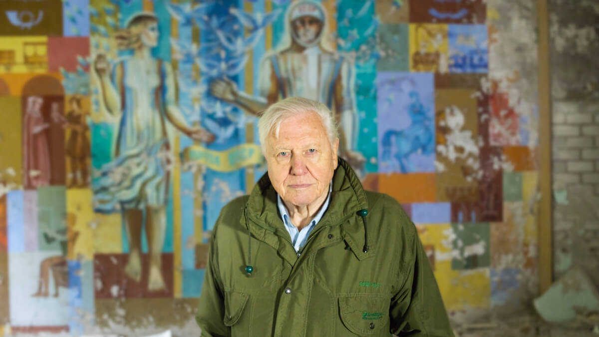 David Attenborough stands in front of a colourful mural which depicts two figures in a renaissance style. Attenborough has a calm yet concerned expression on his face.