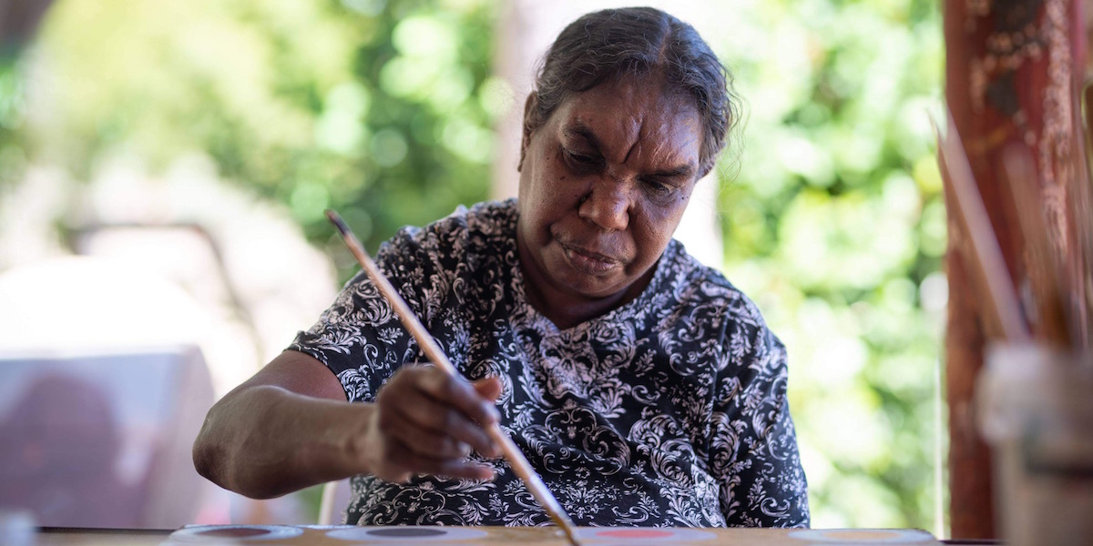 An older indigenous woman faces the camera, she is seated and holds a paintbrush in her left hand and is painting. The background is green and leafy. For Revealed Exhibition 2021