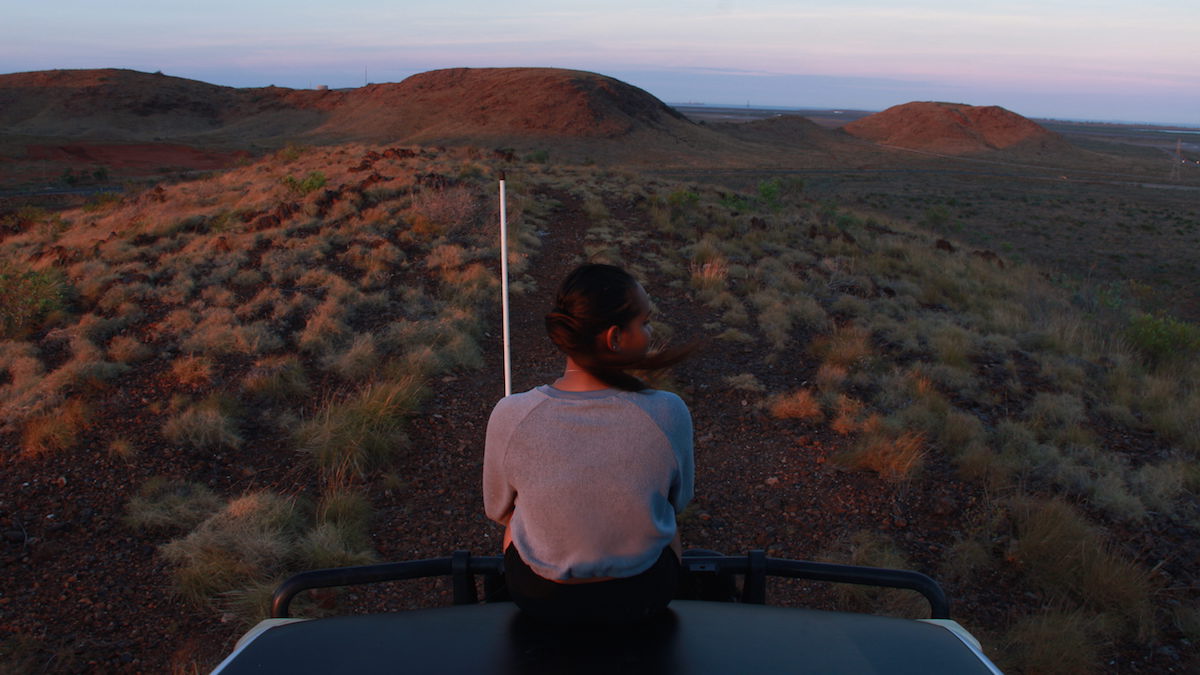 A girl looking into the distance sits on the hood of a 4WD at sunset, looking at the red dirt scrublands and small hills in the distance. She is looking to the right of the image. For Revealed Exhibition 2021