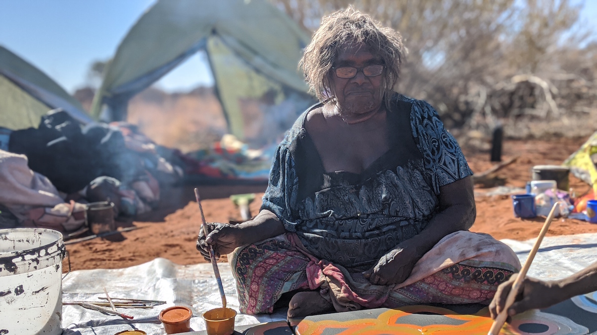 A woman is seated on a tarp, with tents and red dirt in the background. She is painting with ochre colours on a canvas on the ground in front of her. For Revealed Exhibition 2021